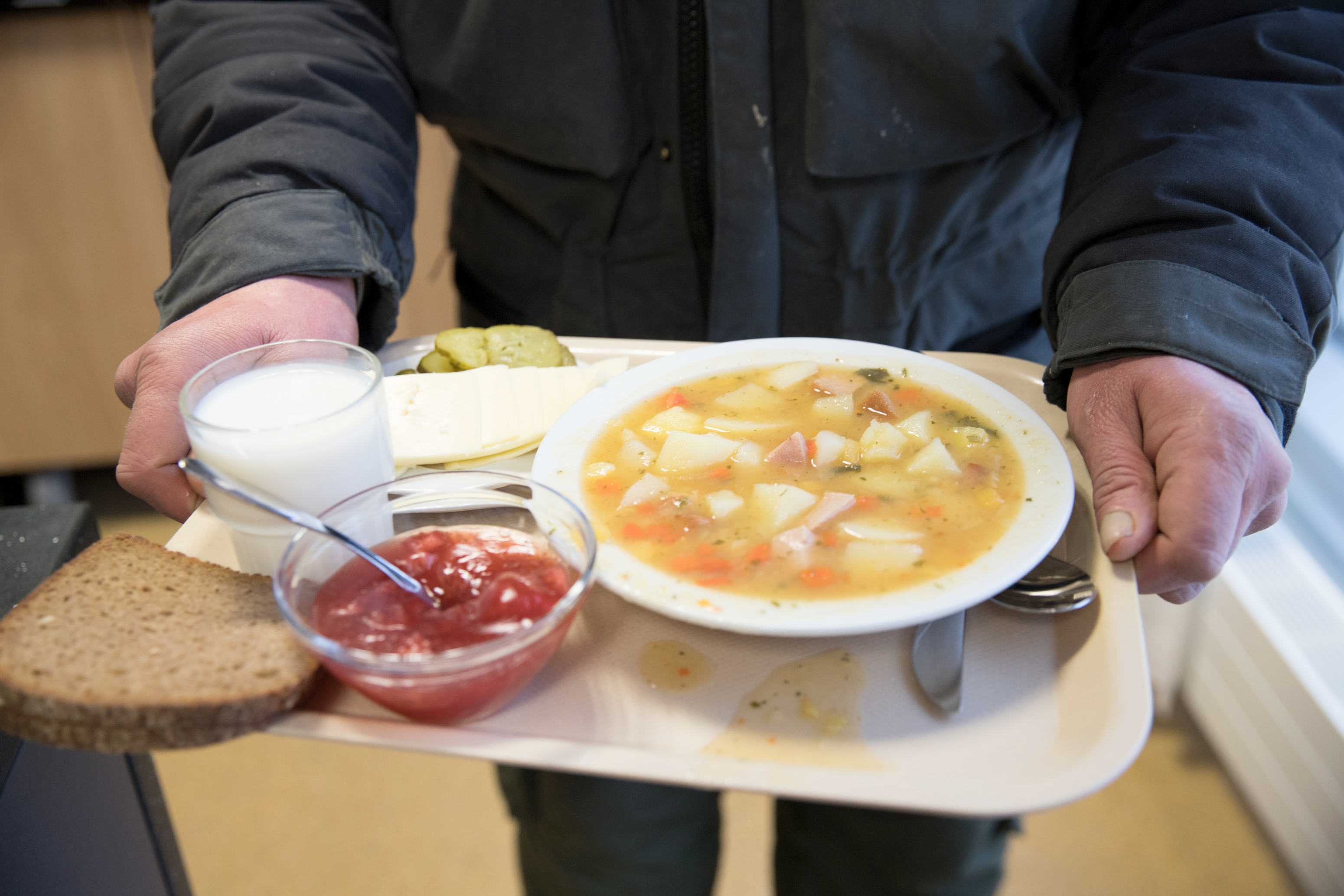 Tarjotinta pitelevät kädet. Tarjottimella ruokalautanen, jossa keittoa sekä leipä, kiisselikulho ja maitolasi