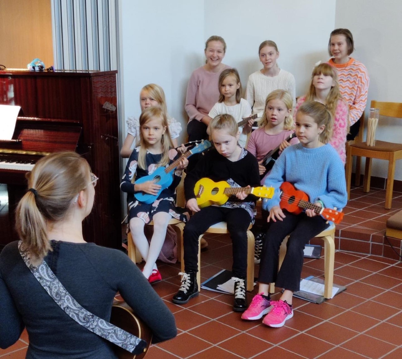 Kuunloiste Kuoronjohtaja on lasten edessä ukulele kädessä. Kuorolaiset katsovat kuoronjohtajaan.