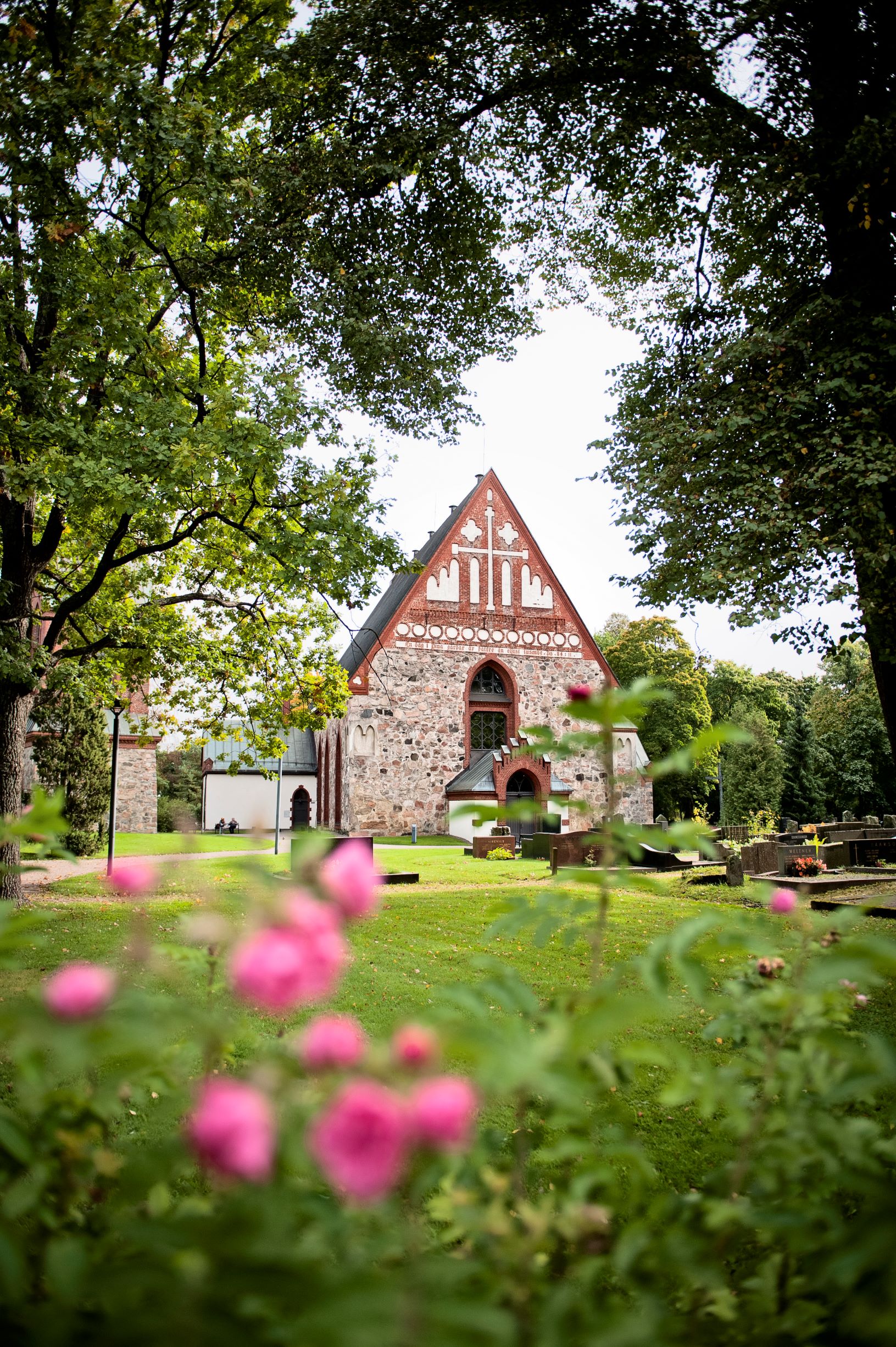Kuvassa Pyhän Laurin kirkko vehreän kirkkomaan ympäröimänä, etualalla kukkia.
