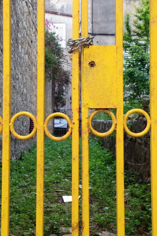 Keltainen portti, joka laitettu kiinni kettingillä. Portin takana näkyy vihreä polku ja kuusi.