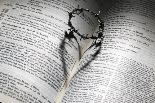 An open Bible with a small crown of thorns on top. The crown casts a shadow shaped like a heart on the Bible.