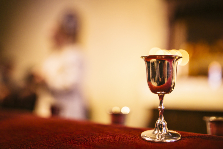 A chalice on the altar. In the background, a priest is sharing the Eucharist.