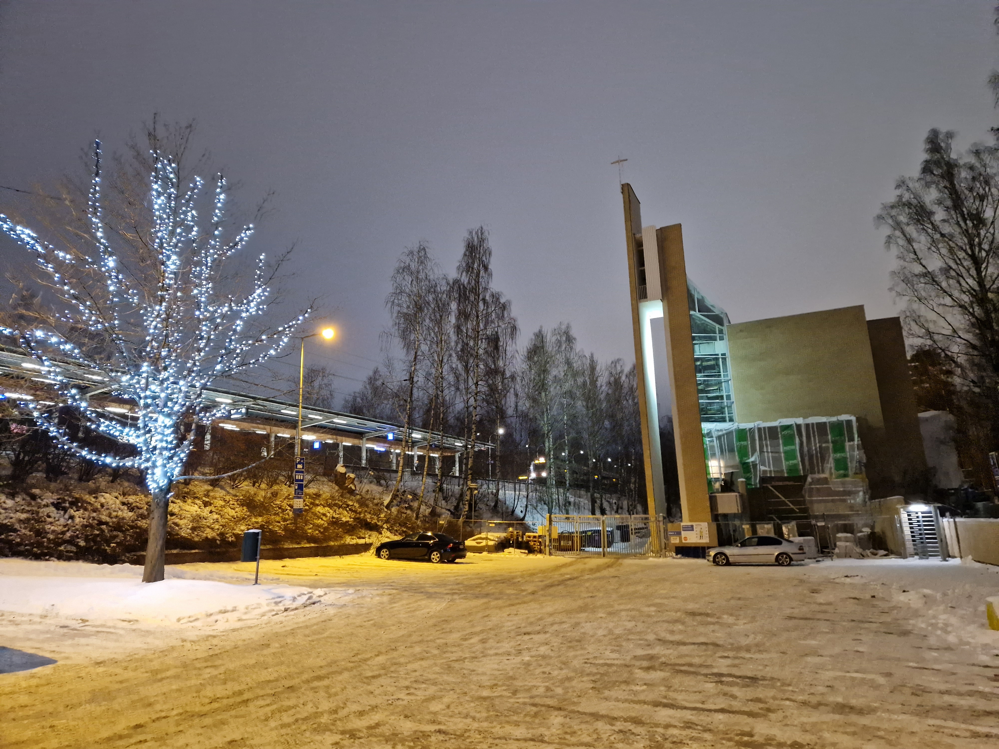 Myyrmäen kirkko talvi-iltana, taustalla junarata, kirkon vieressä valaistu puu