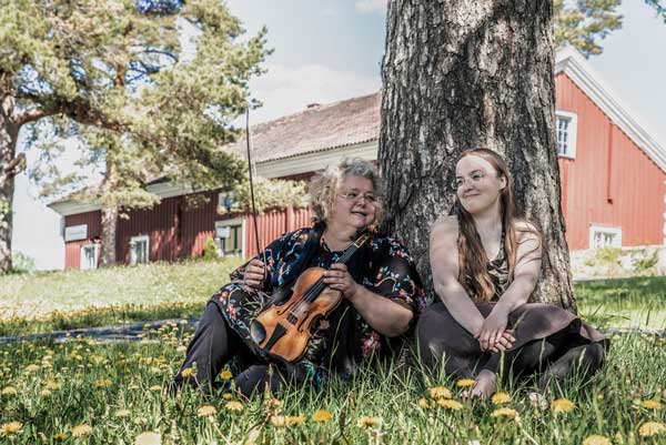 Kreeta-Maria Kentala ja Tea Polso istumassa koivun alla. Kentalalla viulu kädessä.